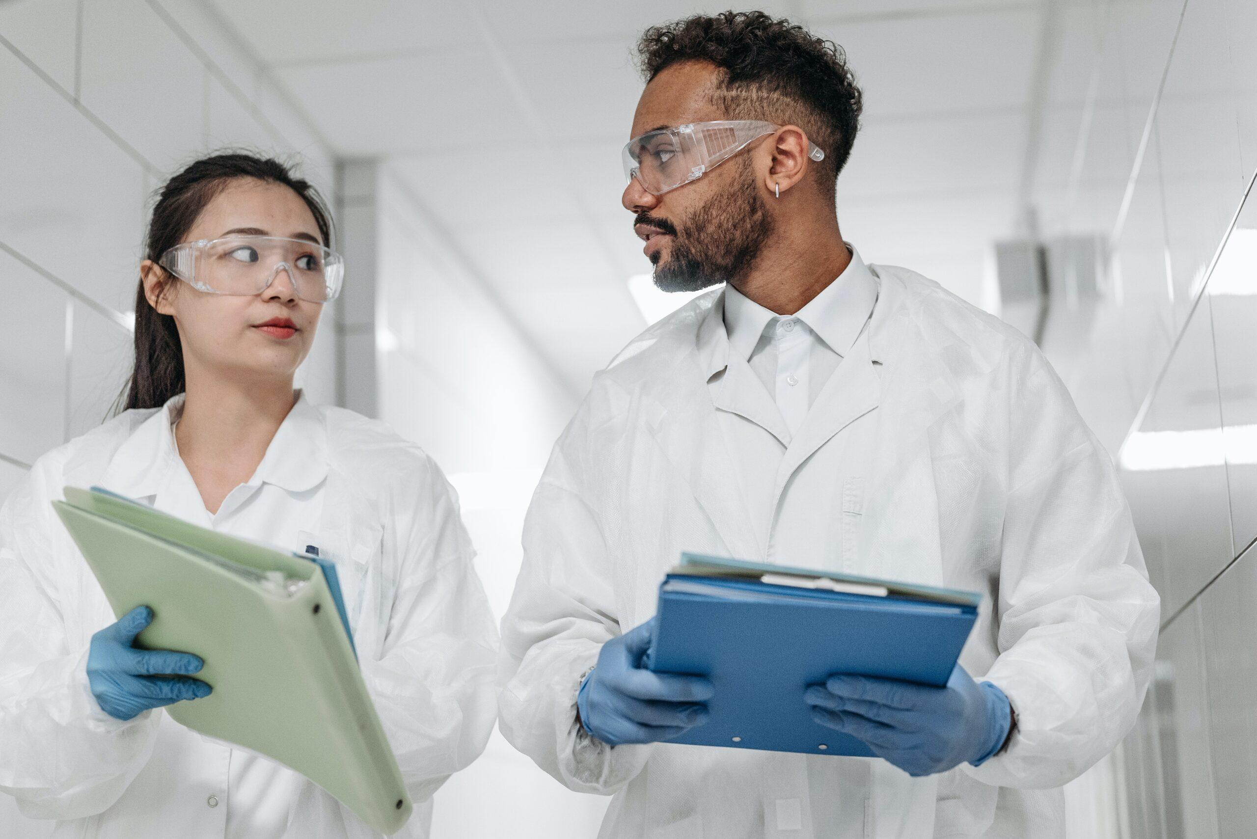two medical lab professionals walking in a hallway wearing personal protective equipment and holding files